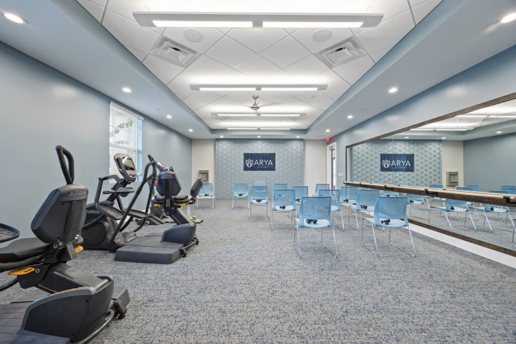 Modern fitness room featuring exercise bikes, large mirror, blue chairs, and ARYA branding on walls.