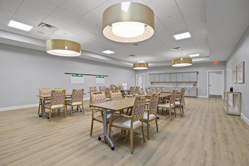 Elegant activity room with wooden tables, patterned chairs, modern lighting, and a kitchenette area.