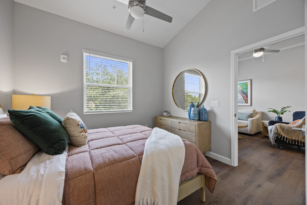 Comfortable bedroom with warm decor, round mirror, soft bedding, and natural light from window.