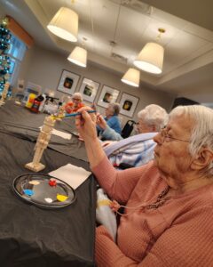 Resident at a senior living community painting a wooden nutcracker figurine during a holiday craft session, with a paint palette featuring bright colors on the table.