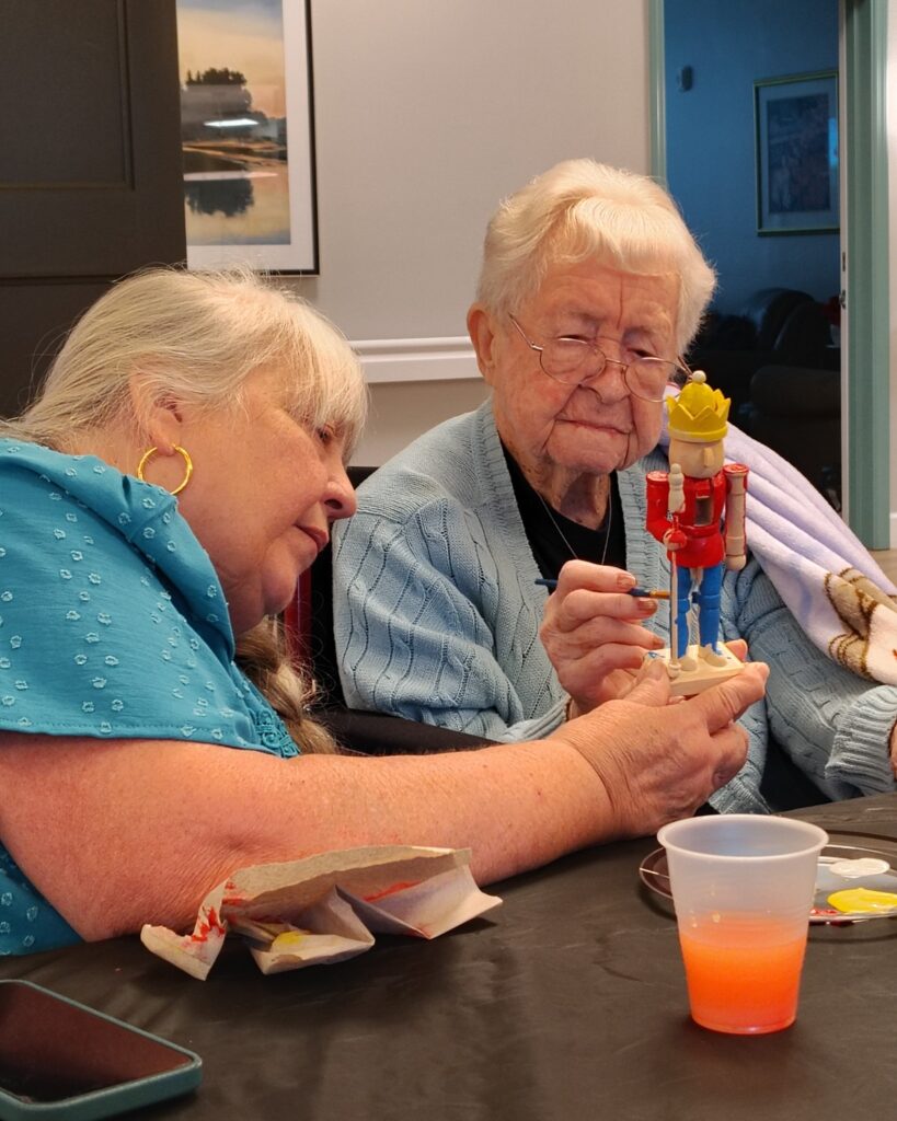 Residents at a senior living community working together to paint a wooden nutcracker figurine during a holiday craft session, with paint supplies and a drink on the table.