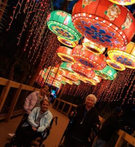 Residents from a senior living community enjoying a vibrant Asian Lantern Festival, walking under colorful illuminated lanterns and sparkling string lights on a wooden pathway at night.
