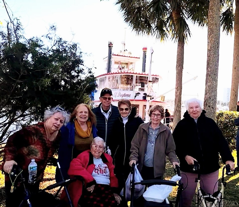 Group of senior living community residents enjoying an outdoor excursion by the Halifax River, posing near palm trees with a classic riverboat in the background.